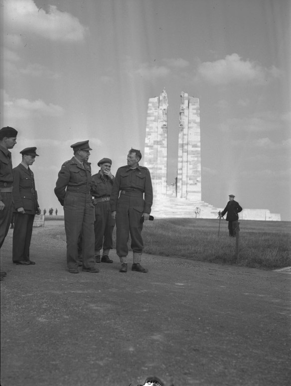A group of men in military uniforms talking, watched from a distance by a man in civilian clothes. A stone war memorial featuring two tall vertical columns can be seen in the background.