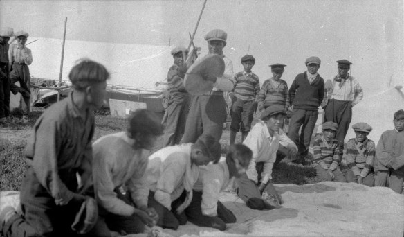 A black-and-white composite photograph of about 16 boys, some standing, some kneeling on the ground. They are playing Dene Handgame. One young man is standing and hitting a caribou-skin hand drum with a wooden drumstick. 