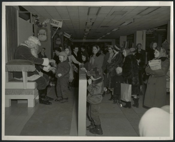 Black-and-white photograph showing a little boy talking with Santa Claus, while other children and many parents, dressed for winter, are lined up nearby. In the background are two signs that read “Trains” and “Meccano.”