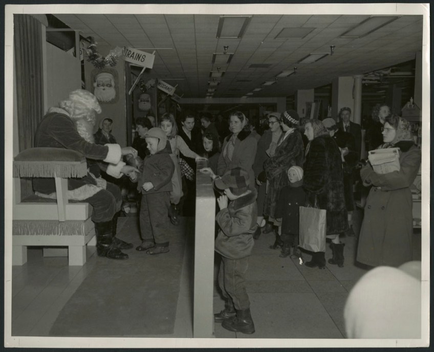Black-and-white photograph showing a little boy talking with Santa Claus, while other children and many parents, dressed for winter, are lined up nearby. In the background are two signs that read “Trains” and “Meccano.”
