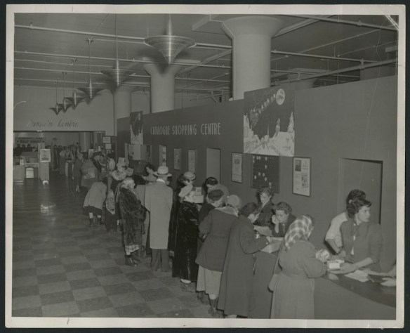 Black-and-white photograph showing shoppers, mostly women, wearing winter coats and hats, studying catalogues at a counter, as well as the store staff behind the counter. There is a sign on the wall reading “Catalogue Shopping Centre,” together with Christmas decorations.