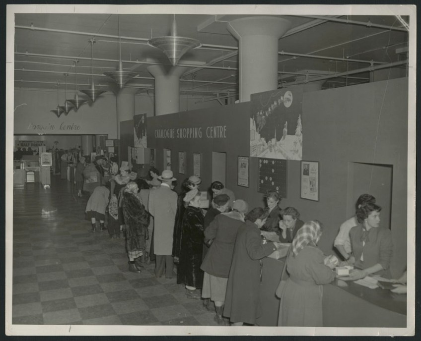Black-and-white photograph showing shoppers, mostly women, wearing winter coats and hats, studying catalogues at a counter, as well as the store staff behind the counter. There is a sign on the wall reading “Catalogue Shopping Centre,” together with Christmas decorations.