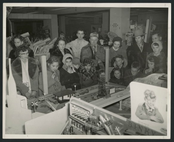 Black-and-white photograph showing a large group of men, women and children watching an electric train in a store.
