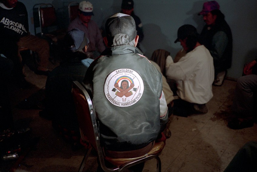 A colour photograph of the back of an elderly man wearing a “Sayisi Dene Traditional Handgame Club” jacket, watching a Dene Handgame match.