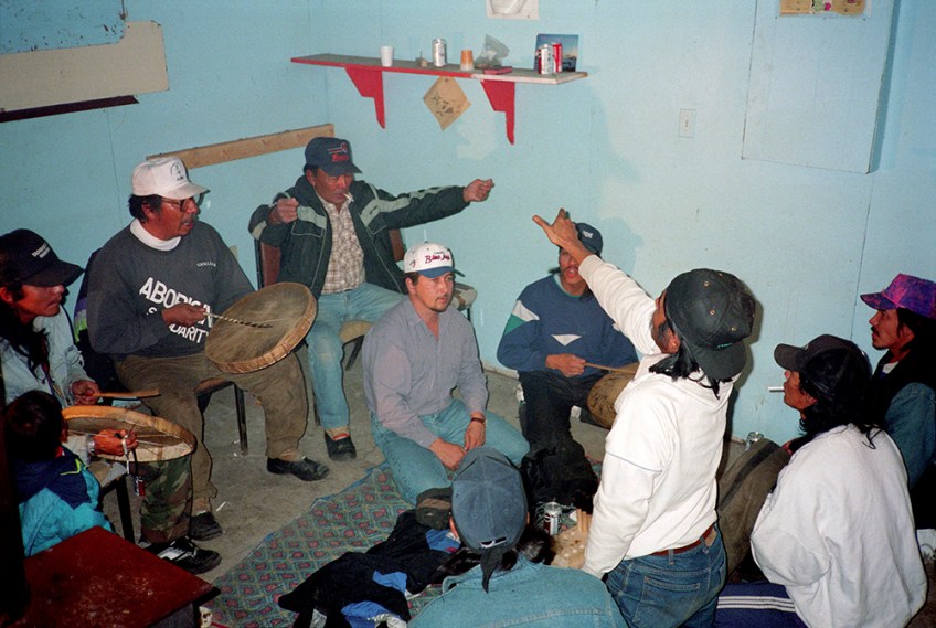 A colour photograph of eight men and one child. The men are playing Dene Handgame. Three of the men are hitting individual caribou-skin hand drums with wooden drumsticks. Two men are gesturing with Dene Handgame hand signals.