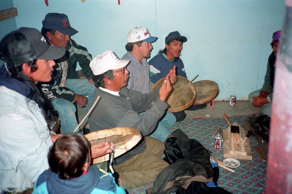 A colour photograph of six men and one small child. The men are playing Dene Handgame. Three of the men are hitting individual caribou-skin hand drums with wooden drumsticks and singing. 