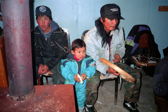 A colour photograph of a man, a teenage boy and a small child watching a Dene Handgame match. The man is hitting a caribou-skin hand drum with a wooden drumstick. The small child is mimicking the drumbeat with his own small hand drum. 
