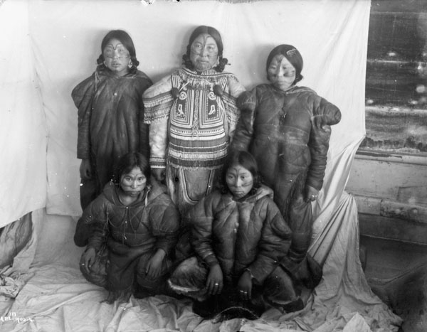 A black-and-white photograph of five Inuit women with facial tattoos standing in front of a white cloth backdrop