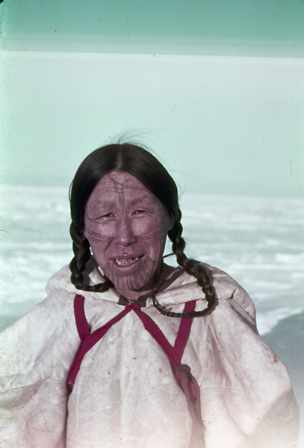 Left: A colour photo of an Inuk woman with facial tattoos wearing a white parka with red straps looking at the camera.