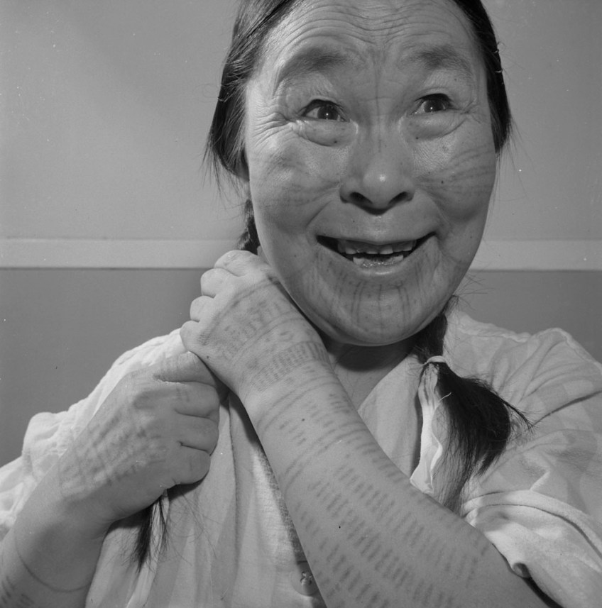 A black-and-white photograph of an Inuk woman with tattoos on her face and arms smiling while braiding her hair.