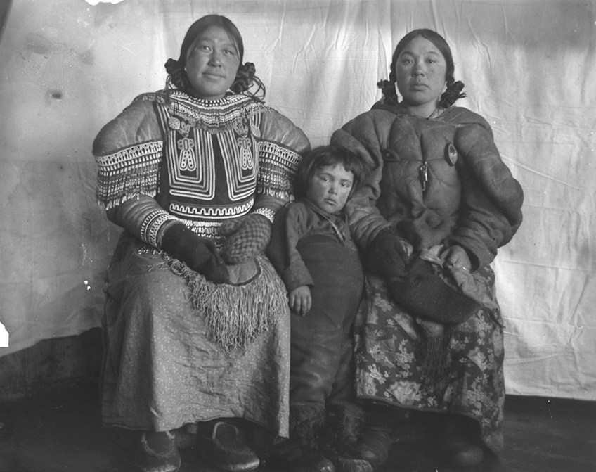 Black-and-white photograph of two women and a child wearing parkas, sitting in front of a fabric background.