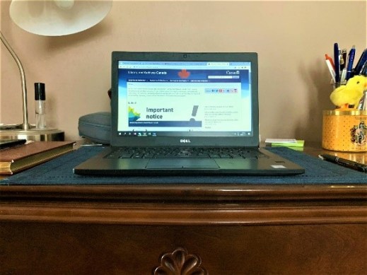 A colour photograph of a laptop on a wooden table, with a lamp and pens in the background.