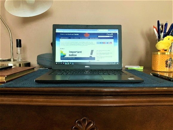 A colour photograph of a laptop on a wooden table, with a lamp and pens in the background.