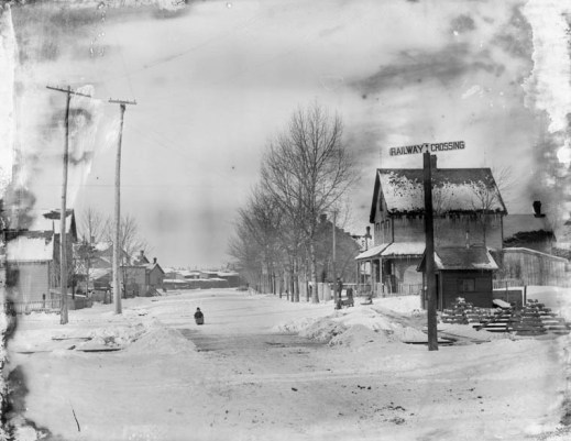 A black-and-white photograph of a street in winter. There is a signpost showing a railway crossing, houses, a person walking in the street and power line poles.