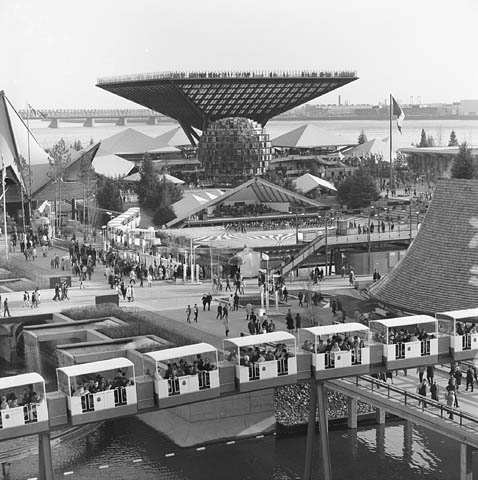 A black-and-white photograph of a crowd of people walking around large pavilions near the waterfront. There is a train with white cars in the foreground. 