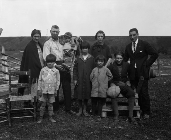 A black-and-white photograph of nine people facing the camera. A man is holding a baby in a cradleboard. Uncut brush landscape is in the background. There are no buildings on the horizon