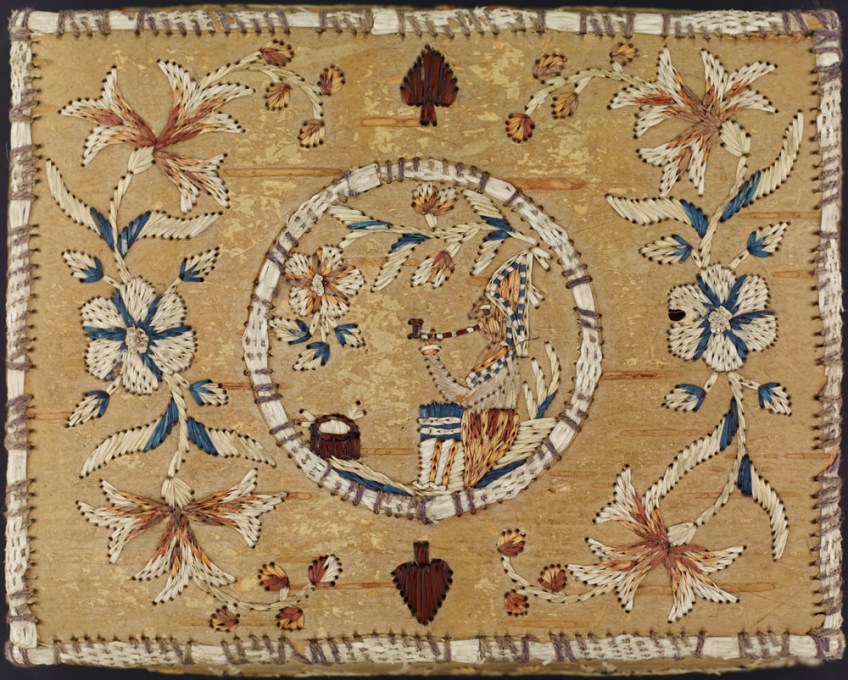 A slightly faded birch bark box lid embroidered with a First Nations figure sitting in profile and smoking a pipe inside a central circle. Red, white, and blue flowers are embroidered on either side of the circle, and a playing card spade is embroidered above and below the circle.