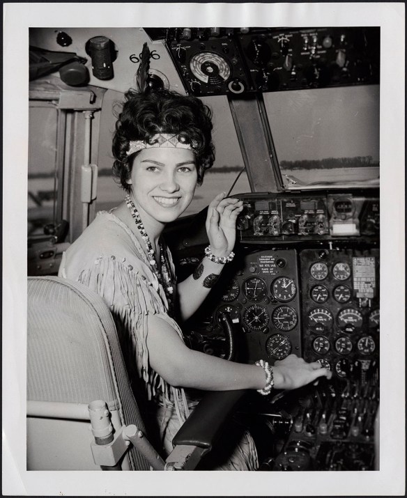 A black-and-white photograph of a woman smiling at the camera wearing buckskin regalia, a necklace, bracelets and a headband.