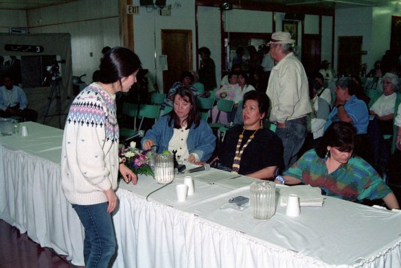 A colour photograph of a woman with a white sweater standing and talking to three women, who are seated on the other side of a long table. 