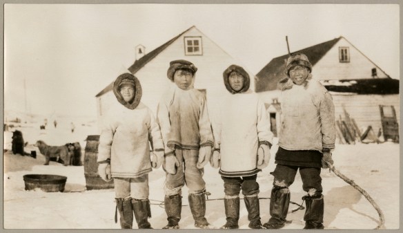 A black and white photograph of four boys in parkas looking towards the camera. There are two buildings in the background. 