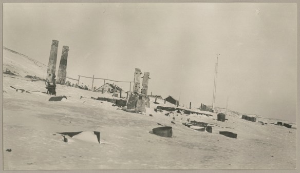 A black and white photograph of the ruins of a building with snow-covered items scattered around. 