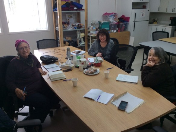 A colour photo of three women smiling at the camera, sitting around a wood boardroom table. 