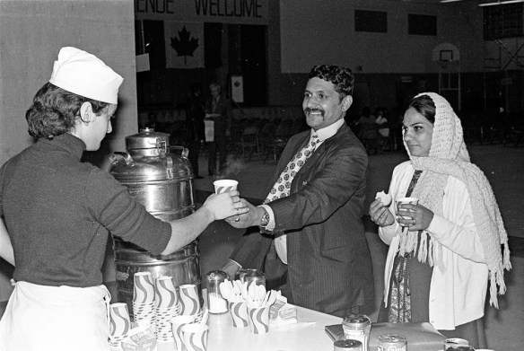 A black-and-white photograph of a woman in an apron and a hat handing a paper cup to a smiling man in a suit, as a woman in a scarf holds a cookie and a paper cup.