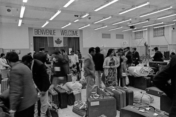 A black-and-white photograph of a large group of people standing in a big room, with luggage and suitcases on the floor, and a Canadian flag and a sign reading “Bienvenue, Welcome.”