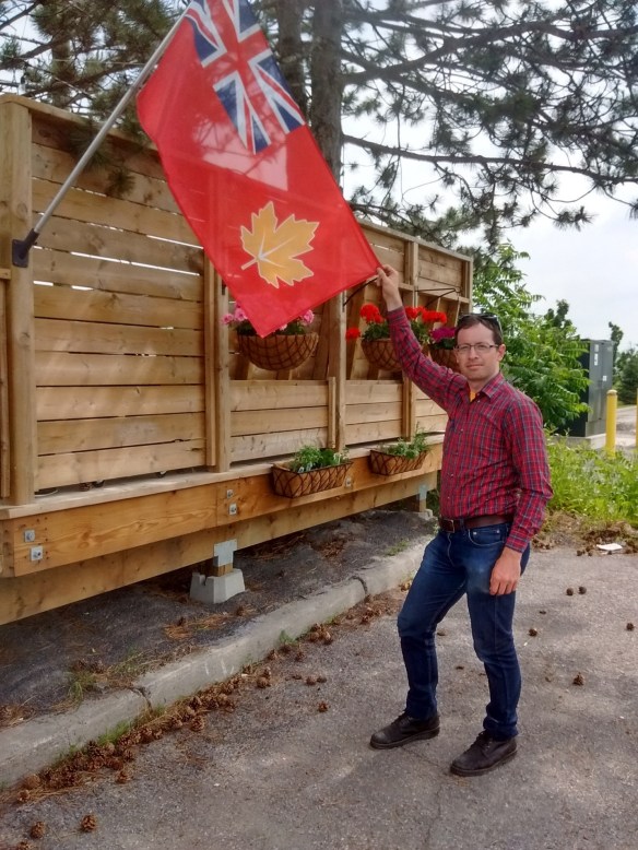 A man standing outside, facing the camera wearing jeans and a red plaid shirt holding the corner of a flag.