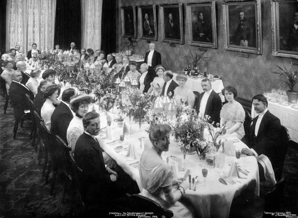 Black-and-white photo of people in formal attire seated around a long oval table. The table features place settings and decorative centrepieces.
