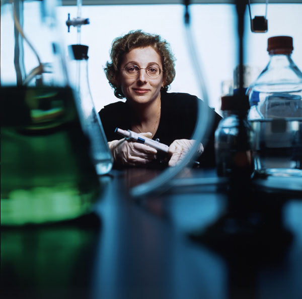 Colour photograph of a woman with glasses looking towards the camera. Beakers and bottles containing liquids are in the foreground.