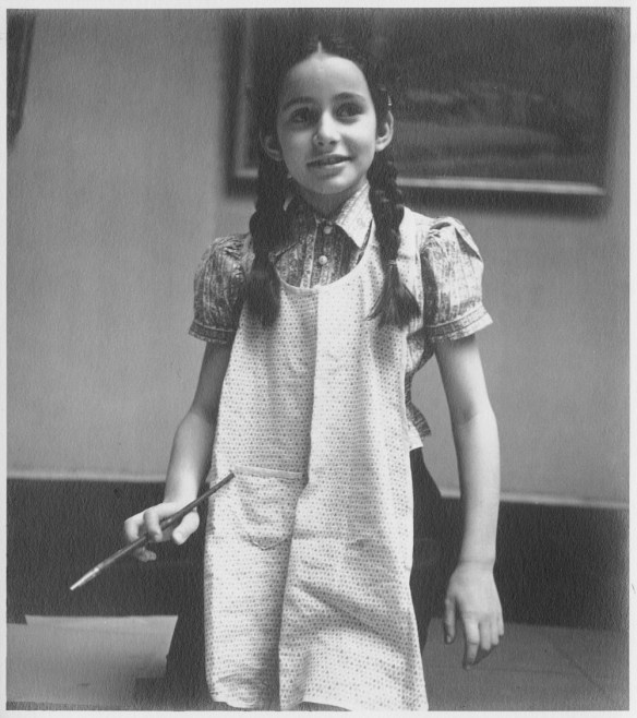 A black-and-white photograph of a girl with dark braids and a light apron kneeling on the floor and holding a paintbrush in her right hand. The bottom of a framed painting can be seen behind her.