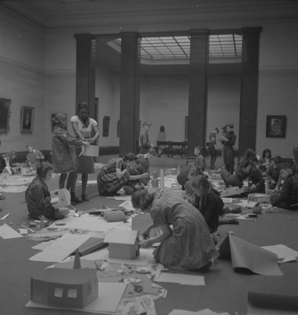 A black-and-white photograph of children kneeling in the middle of the floor in a gallery, surrounded by paper and art supplies. A teacher stands near the middle of the room, assisting a student. The walls are hung with framed paintings, and an adjacent gallery is visible behind four dark columns. The scene is full of energy as the children build paper houses.