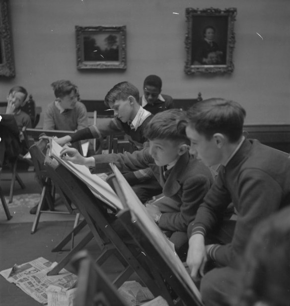 A black-and-white photograph of six boys sitting on chairs in a gallery. Each boy has a second chair in front of him being used as a drawing easel. Two framed paintings can be seen on the wall in the background, and there are newspapers scattered on the floor.