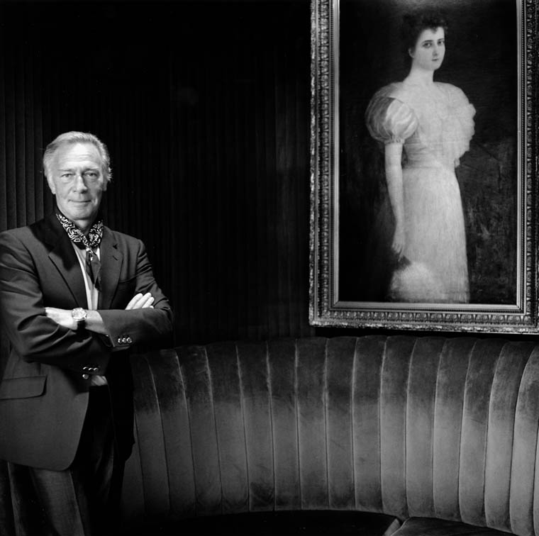 A black-and-white photograph of Christopher Plummer in a suit standing on the left with his arms crossed. A large framed painting of a woman in a dress holding a fan is hung on the right side.