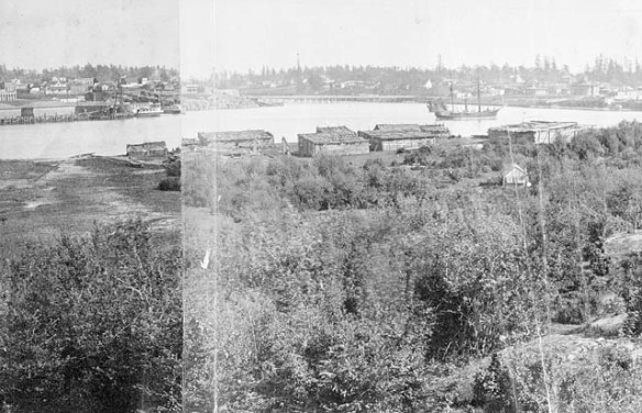 A black-and-white photograph of villages on either side of a harbour, with a large ship on the water. There are forests behind each village. 