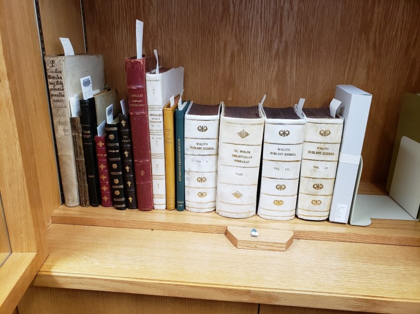 A colour photo of book with different coloured spines on a wooden shelf. The books have small white pieces of paper sticking out of their tops.
