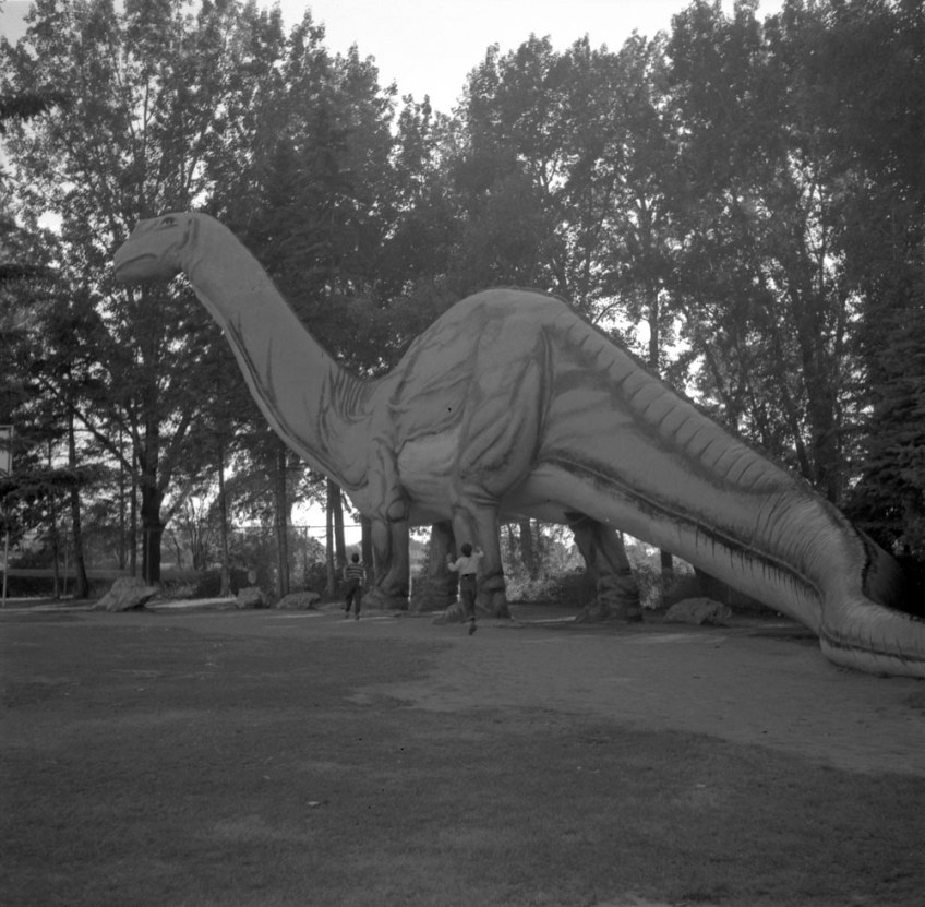 A large sculpture of a brontosaurus in front of some tall trees, with two children running toward it.