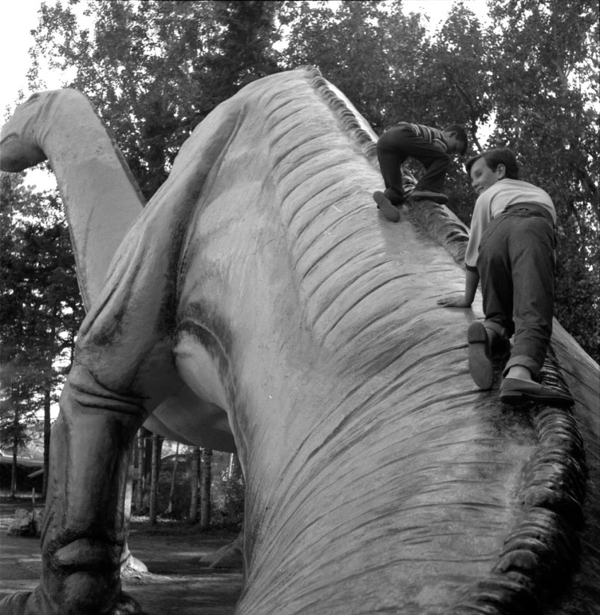 A black-and-white photograph of two children climbing up a large model dinosaur.