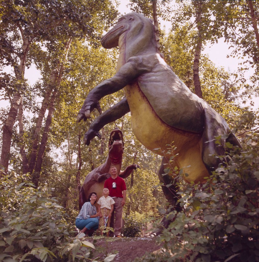 A woman, child and man stand under a large model of a dinosaur, surrounded by trees. The group is looking toward another dinosaur model.