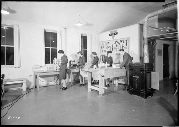 A black-and-white photo of four women and a man in a shop with tools and tables. There are three windows and a sign that reads YMCA.