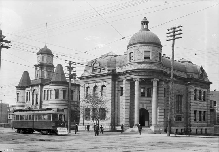 A black-and-white photograph of two ornate buildings, one with a columned portico and a cupola. People walking, a street car, and power lines are in the foreground.