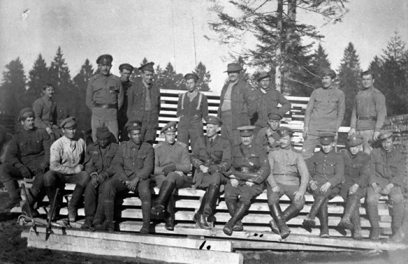 A black-and-white photograph depicting 21 soldiers. They are casually posed outdoors, either sitting or standing on a pile of long wooden planks. Eight of the men appear to be in Russian uniforms, and the rest are Canadians, including two Black men.