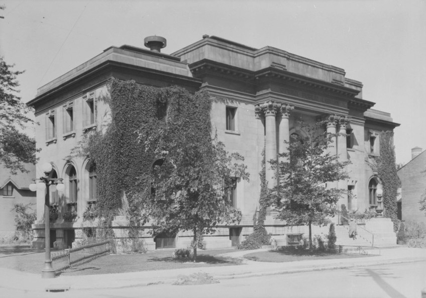 A black-and-white photograph of a two-storey stone building with a columned portico and ivy growing up its sides.