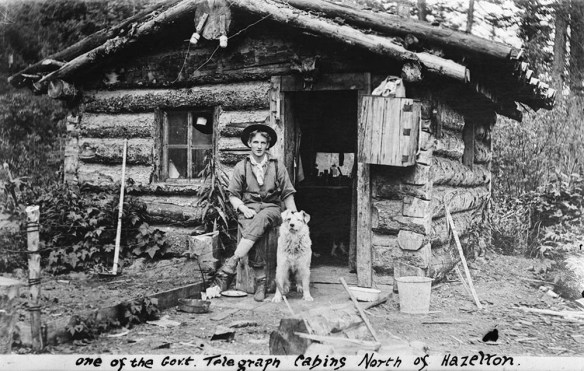 Black-and-white photograph of a young man and his dog sitting in front of a one-room log cabin.