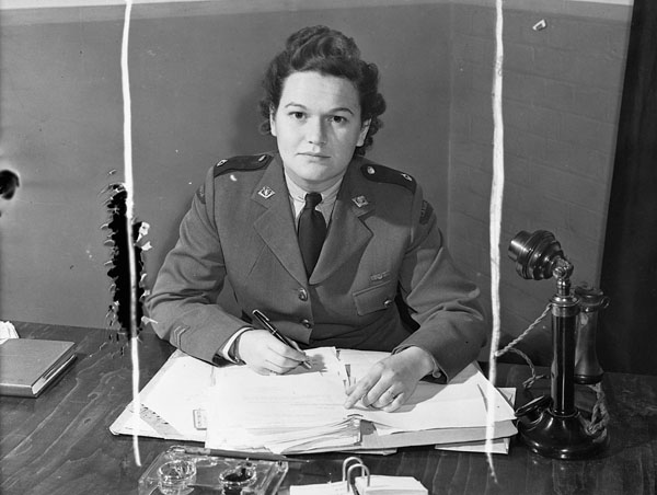 A black-and-white photograph of a woman in a military uniform looking at the camera. She has a pen in her right hand, papers on her desk and a black candlestick-style telephone to her left.