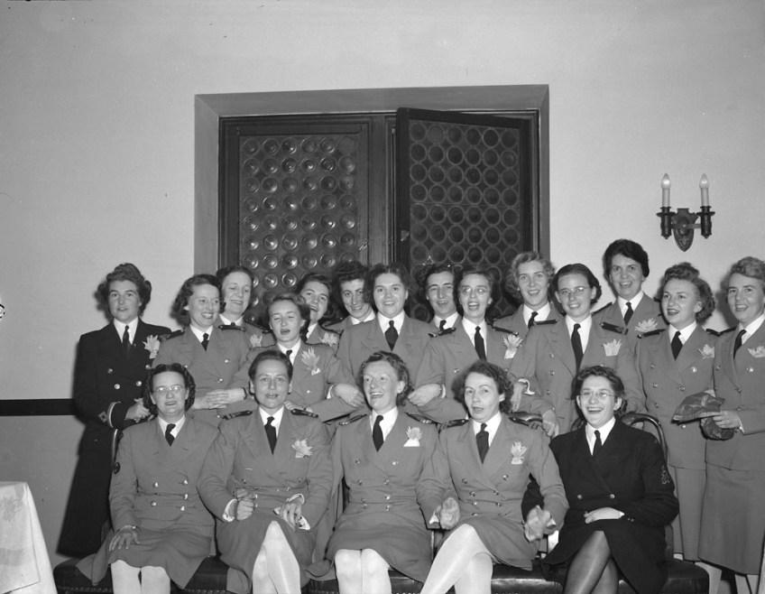 A black-and-white photo of a group of women in military uniforms smiling at the camera. There are two women in dark suits. The women in the first row are seated and holding hands. Some of the women standing in the back row have their arms linked.