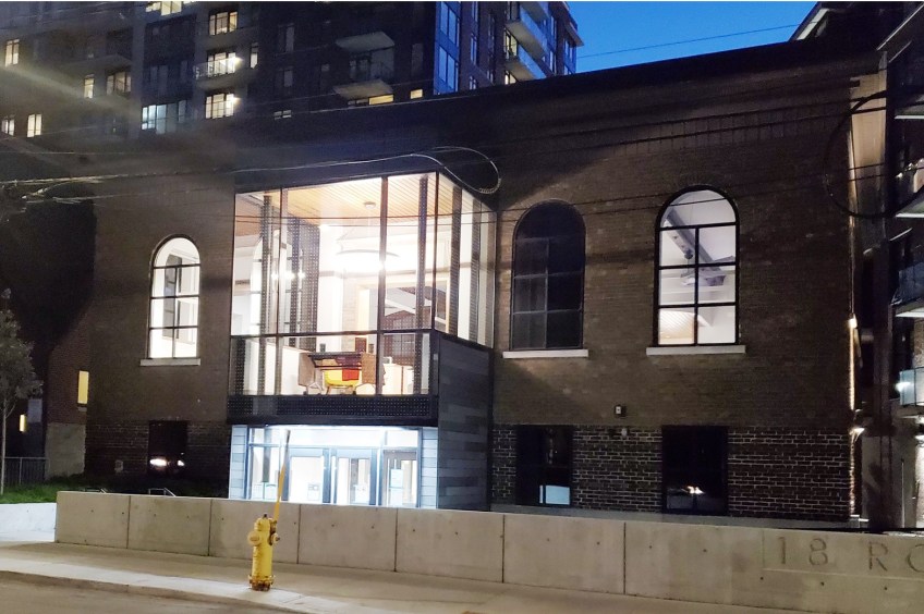 A brown brick 2 storey building with curved windows on the top floor. The entrance is glass and there is a yellow fire hydrant in the foreground.