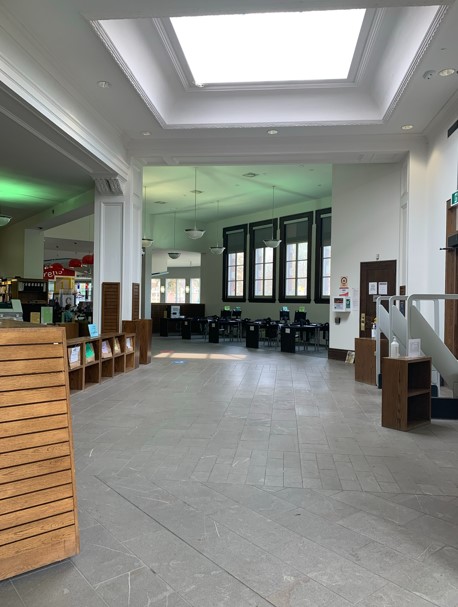 A colour photograph of the inside of a library. A large skylight, book shelving and computer terminals can be seen in the room. There are four windows at back.