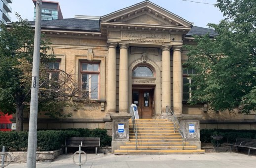 A colour photograph of a beige building with a columned entrance and a pediment above the front door. There is single set of stairs leading to the building. The words “Public Library” are etched above the entrance.